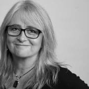 Woman with long hair looks directly at camera smiling warmly, wearing black rimmed glasses.